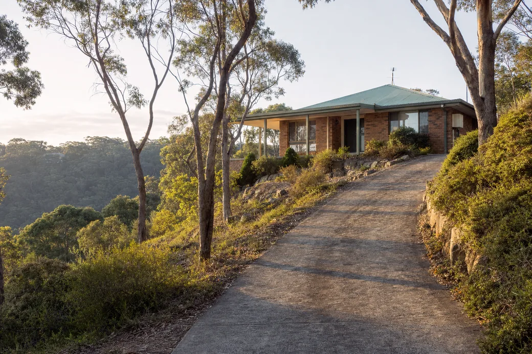 A brick-veneer house with a green iron roof at the top of a long sloping driveway, gums and a bushland ridge behind in late sun — pressure cleaning, Central Coast. Illustrative example.