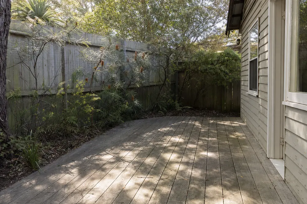 A weathered grey timber deck and paling fence behind a weatherboard cottage, banksia flowering along the fence — deck and fence cleaning, Central Coast. Illustrative example.