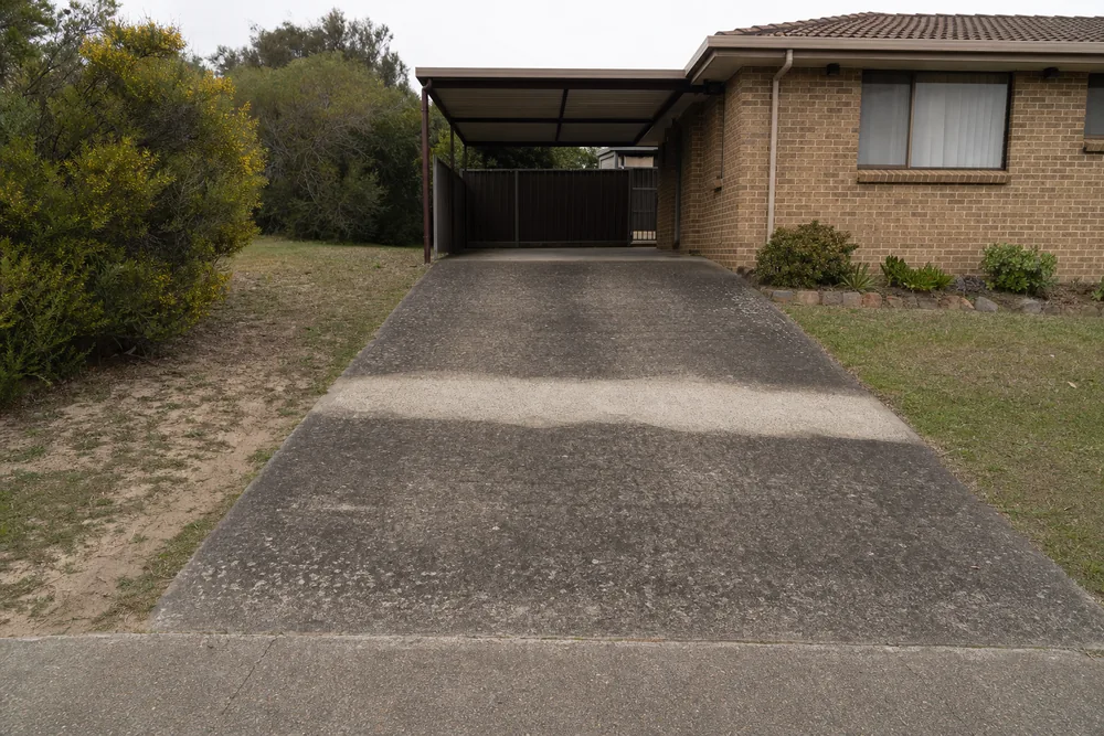 A grimy concrete driveway leading up to an empty carport beside a brick-veneer house, one uneven strip across it cleaned back to pale grey — driveway cleaning, Central Coast. Illustrative example.